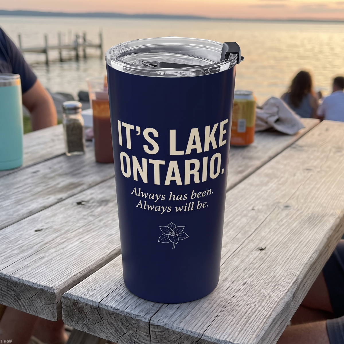 A navy blue insulated tumbler with a clear lid sits on a weathered wooden dock table outdoors. The tumbler is printed with the text 'IT'S LAKE ONTARIO.